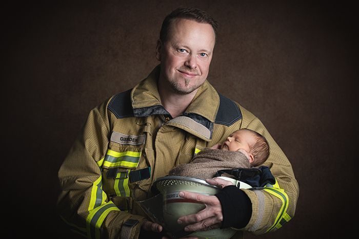 Babyfotografie mit Vater im Studio in Düsseldorf – Neugeborenes liegt geborgen im Arm seines Vaters in Feuerwehrkleidung